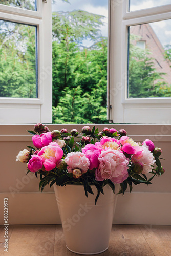 Big fresh pink and white peony blooming bouquet in a white metal bucket next to white window on a green blurred tree and blue sky background. Spring home decor or present idea. Vivid summer colors.