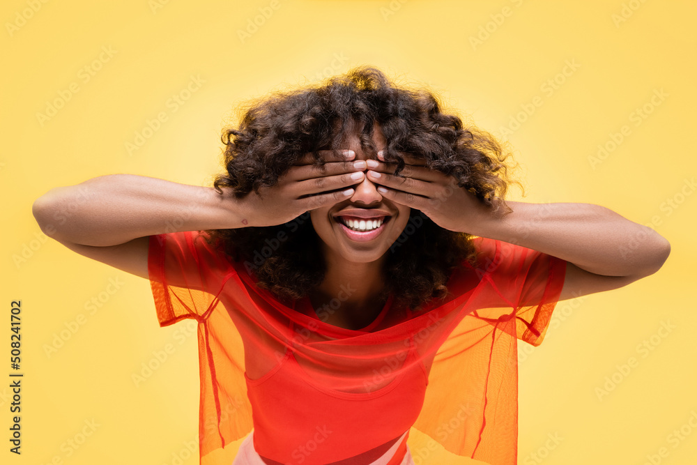 happy african american woman in summer top covering eyes with hands isolated on yellow. happy african american woman in summer top covering eyes with hands isolated on yellow.