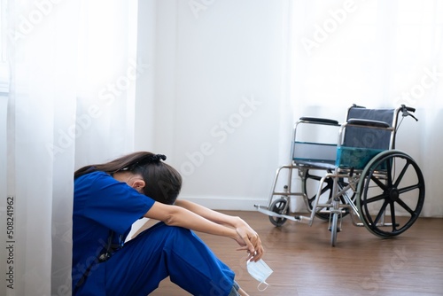 Tired exhausted female asian scrub nurse wears face mask blue uniform sits on hospital floor. Depressed sad Thai ethic doctor feels fatigue burnout stress, lack of sleep, napping at work.