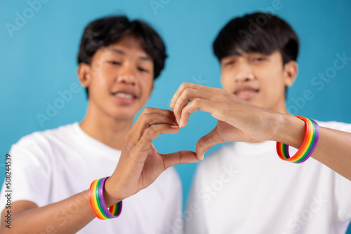  Close up of Asian male couple with gay pride rainbow awareness wristbands showing heart gesture in studio on blue background. lgbt, same-sex love and homosexual relationships concept