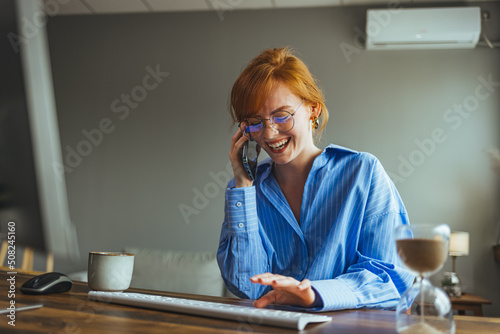 Smiling female employee multitask working on computer in office talking over phone, happy woman worker using laptop, laughing speaking with friend on cell or communicating with client online