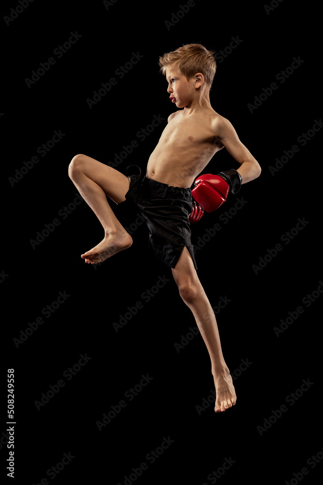 Studio shot of preschool boy, cute kid in sports shots and gloves training alone on black background. Sport, education, action, motion concept.