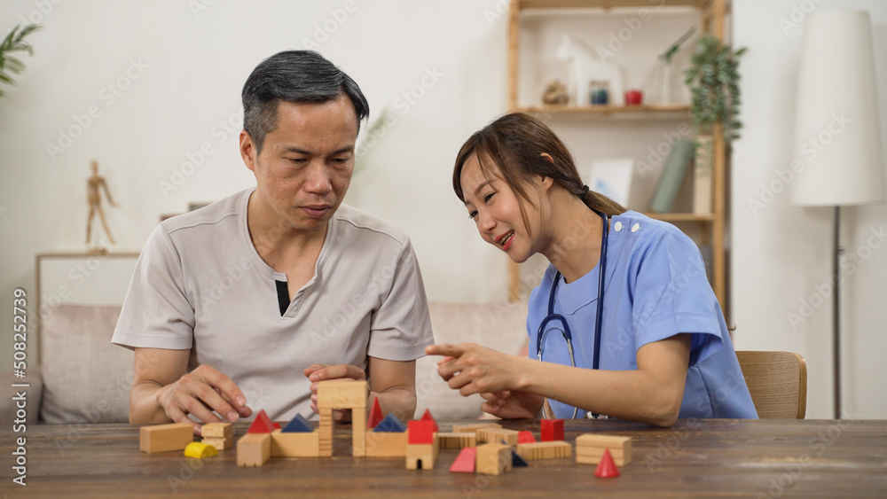 Fototapeta premium asian older male Parkinson’s patient trying to build wood brick with hand tremor while having rehabilitation at home. female nursing aide giving praise with thumb up sign