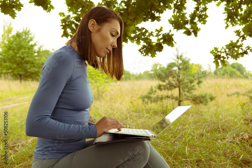 A young Asian woman sits under a tree with a laptop in a field. Freelancer woman brunette running in nature