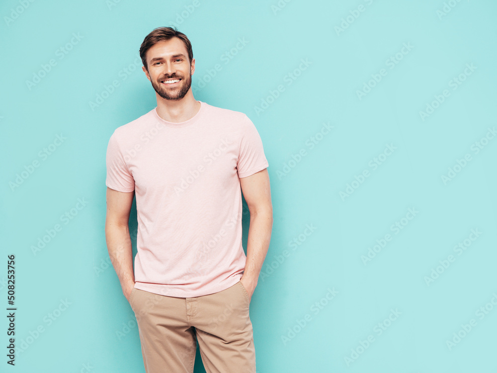 Portrait of handsome smiling stylish hipster lambersexual model. Sexy man dressed in pink T-shirt and trousers. Fashion male isolated on blue wall in studio Portrait of handsome smiling stylish hipster lambersexual model. Sexy man dressed in pink T-shirt and trousers. Fashion male isolated on blue wall in studio