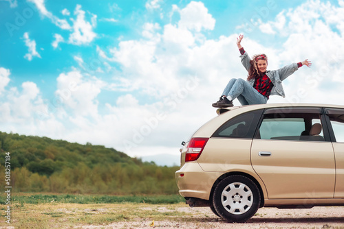 Car trip. Happy teen girl is sitting on the roof of a car, joyfully raising her hands up. Mountains and sky are in the background. Copy space. The concept of freedom and obtaining a driver's license © _KUBE_