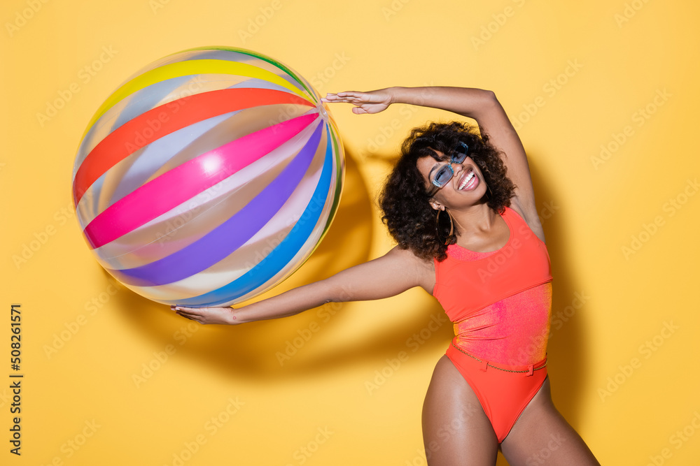 excited african american woman in swimsuit and sunglasses posing with striped inflatable ball on yellow. excited african american woman in swimsuit and sunglasses posing with striped inflatable ball on yellow.