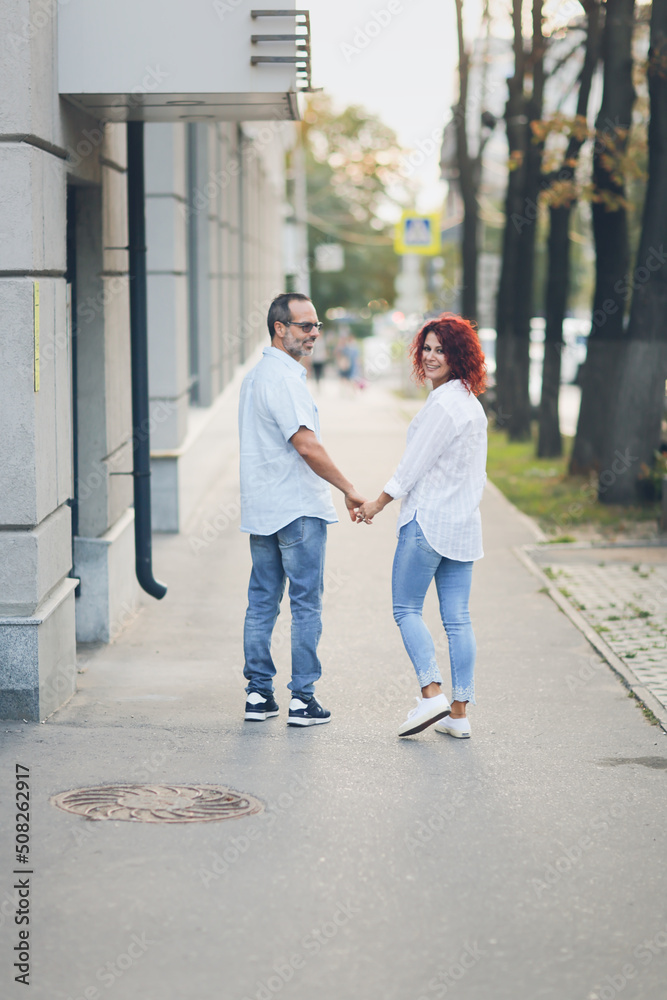 Cute European middle-aged couple holding hands walking in the city street, man and woman walking down the street in summer, back view Cute European middle-aged couple holding hands walking in the city street, man and woman walking down the street in summer, back view