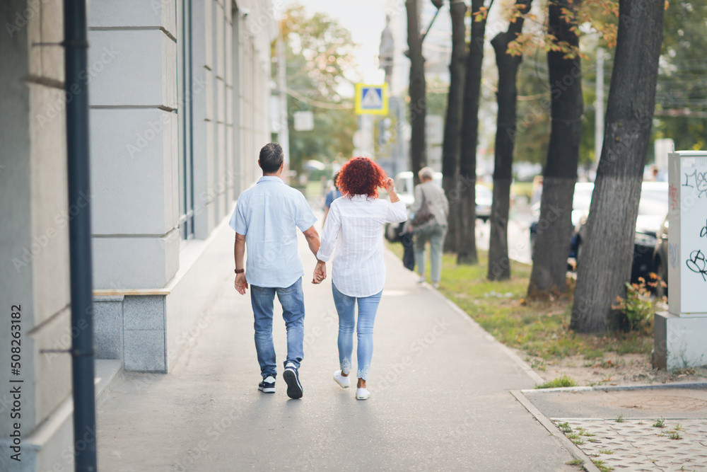 Cute European middle-aged couple holding hands walking in the city street, man and woman walking down the street in summer, back view Cute European middle-aged couple holding hands walking in the city street, man and woman walking down the street in summer, back view