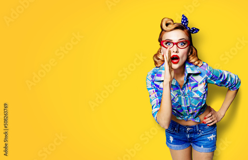 Portrait of beautiful woman in red glasses, holding hand near opened mouth and saying. Pin up girl. Foxy blonde haired pin up girl at retro vintage studio concept, isolated over yellow.