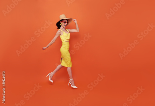 Happy attractive woman in summer dress and hat walking to isolated on copy space orange background.