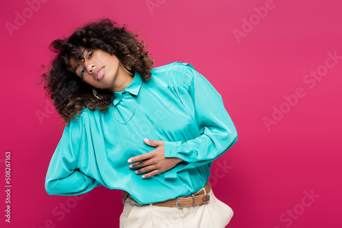 african american woman in turquoise blouse posing with hand behind back isolated on pink. © LIGHTFIELD STUDIOS