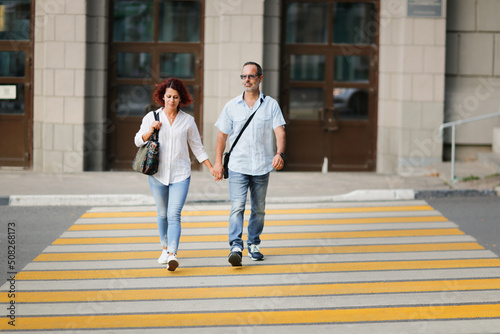 Cute middle aged european couple holding hands and crossing the road on a pedestrian crossing in the cities, summer walks and travel