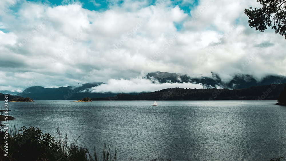 Barco velero navegando un lago calmo con nubes bajas y montañas de fondo. Villa La Angostura, Argentina