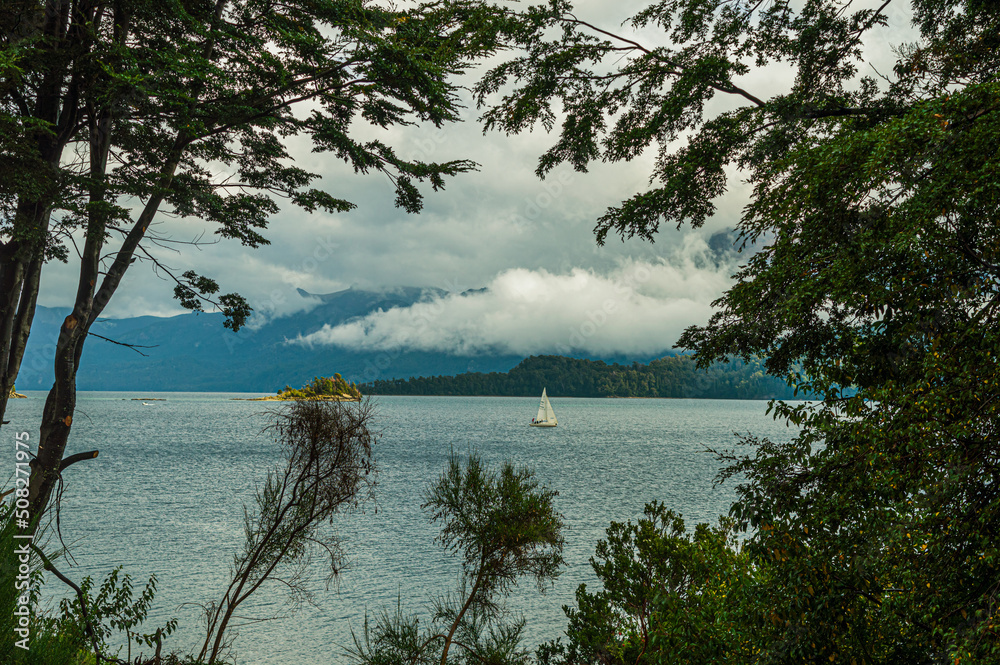 Sailboat navigating a calm lake with low clouds and mountains in the background. View between the trees. Villa La Angostura, Argentina