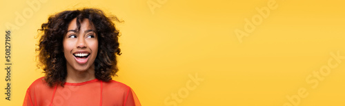 astonished african american woman with curly hair looking aside isolated on yellow, banner.