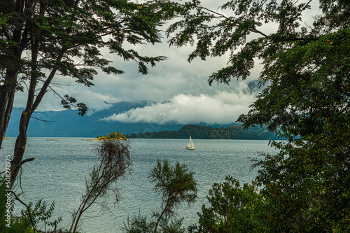 Obraz na płótnie Sailboat navigating a calm lake with low clouds and mountains in the background. View between the trees. Villa La Angostura, Argentina w salonie