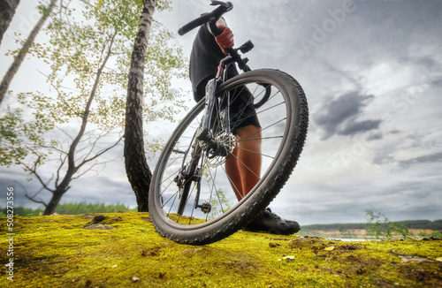 Fototapeta Naklejka Na Ścianę i Meble -  Cyclist stands with a bicycle on a rock overgrown with moss. Low angle view. Sport and travel outdoors.