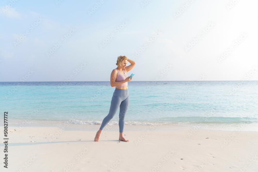 A sporty woman on the beach wearing headphones uses a smartphone.