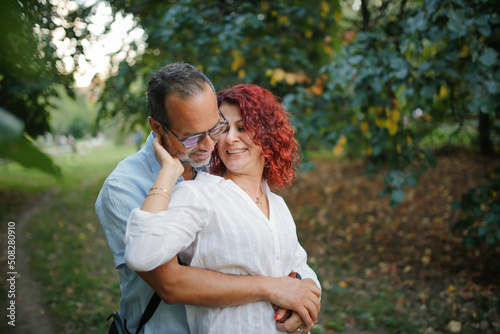 slender man with beard kisses and hugs pretty red-haired curly woman. Cute middle aged european couple hugging in the park