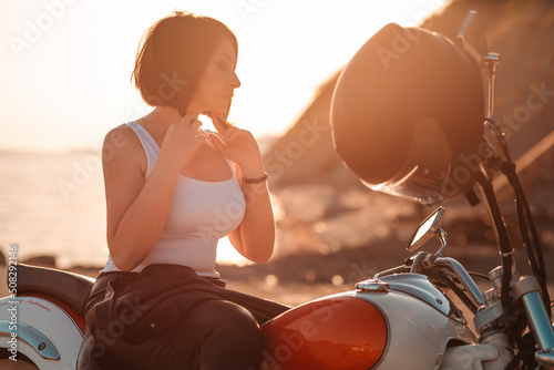 Fototapeta Naklejka Na Ścianę i Meble -  Silhouette of beautiful adult woman standing near motorcycle with helmet. Sunset on the background. Concept of motorcycle travel and feminism