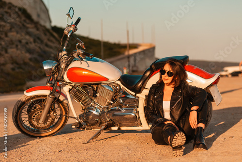 Cool confident woman in a leather motorcycle outfit sitting on the ground near a motorcycle. Sunlight. The concept of the Motorcyclist Day