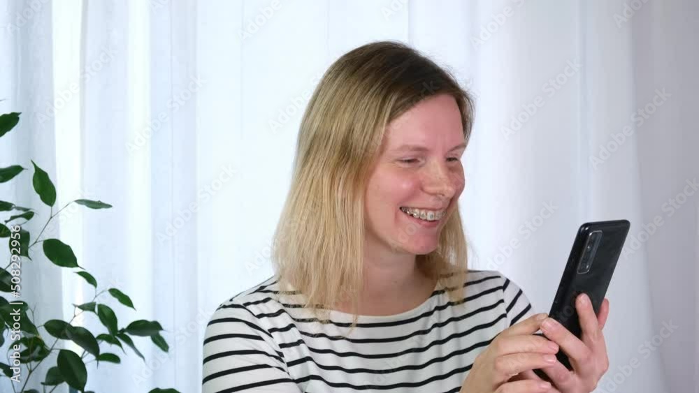 Young smiling caucasian woman with dental braces in casual striped t-shirt making video call on mobile phone. Light background, Beautiful blonde girl with braces on teeth talking by smartphone at home Young smiling caucasian woman with dental braces in casual striped t-shirt making video call on mobile phone. Light background, Beautiful blonde girl with braces on teeth talking by smartphone at home
