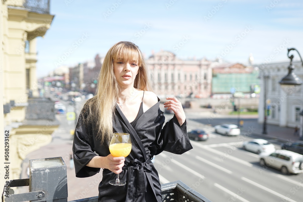 Fototapeta premium In the afternoon, in the light of the sun, a touching girl in black pajamas enjoys a glass of orange juice. She's good on the balcony.