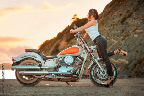 Beautiful sexy woman with a high heels and leather pants, posing confidently with motorcycle. Sunset sky on the background. The concept of Motorcyclist Day © _KUBE_