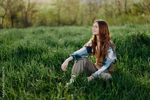 A young beautiful woman sits on the green grass in the park and looks out into the setting sun