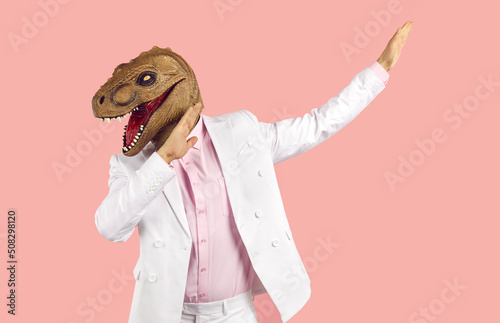 Funny weird guy in wacky animal mask having fun at crazy party. Eccentric man in white suit and silly ugly masquerade dinosaur mask dancing isolated on pink background