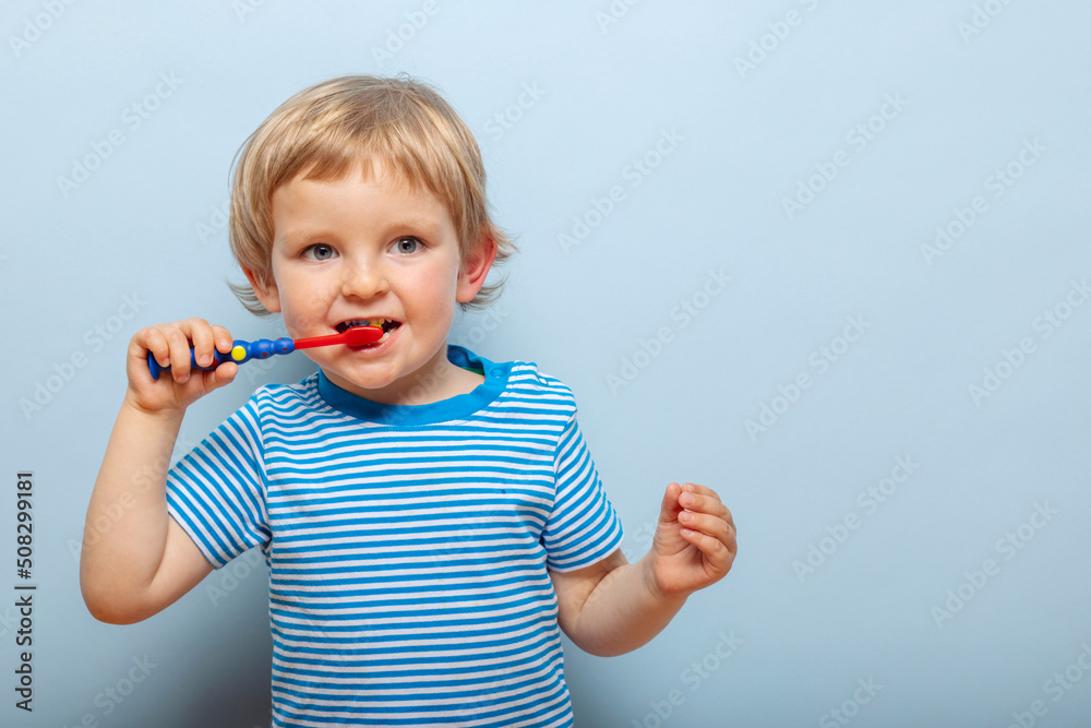 Little blonde boy brushing teeth with toothbrush on blue background. Dental hygiene