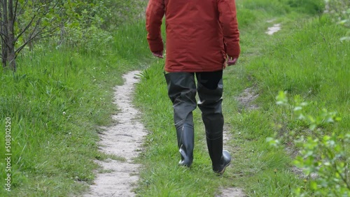 Man in orange parka and rubber boots waders walking in countryside. Path with grass. Back view. photo