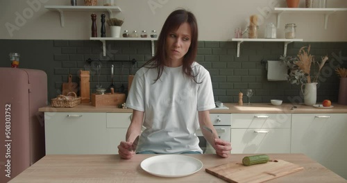 Woman sitting at table, looking sad and bored with diet not wanting to eat slice of cucumber. Healthy nutrition and dieting eating