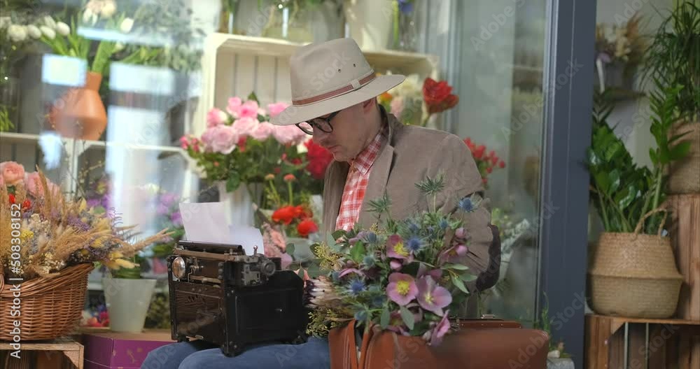 Poet or writer working concept. Cute male person in eyeglasses and hat using old typewriter in flower shop. Suitcase with flower bouquet standing near. 4k video footage Poet or writer working concept. Cute male person in eyeglasses and hat using old typewriter in flower shop. Suitcase with flower bouquet standing near. 4k video footage