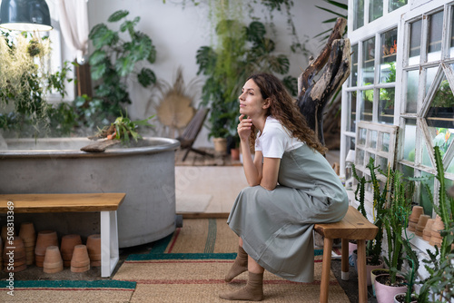 Calm woman sits on bench in home garden surrounded plants thinking, looking out the window. Thoughtful gardener female resting after work enjoying her greenhouse. Doubtful italian lady in floral shop