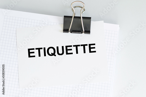 A white card with the text ETIQUETTE on a clip to a white sheet on a light table, business concept