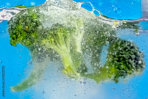 Fresh broccoli slices in water with air bubbles on blue background. Boiling water with fresh vegetables, close-up