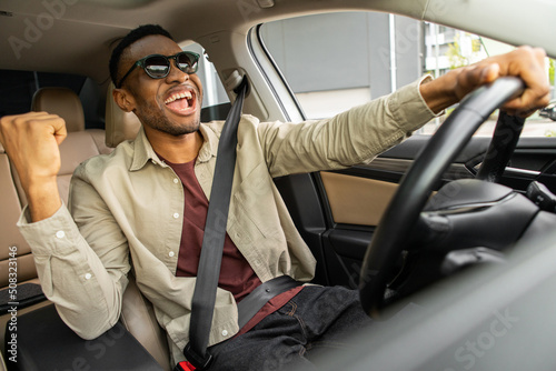 A joyful African American in glasses dances in a car, sings while driving his car Fototapet