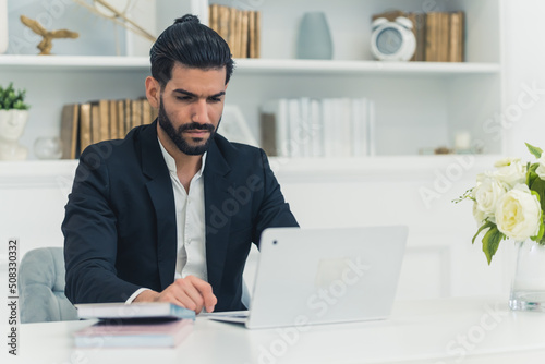 Young businessman sitting at a table at home working on a laptop wearing a black suit. High quality photo