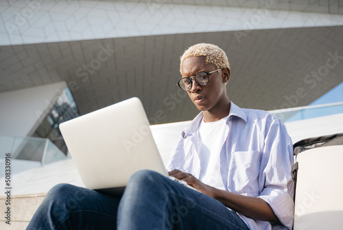 African American programmer using laptop computer working freelance project sitting at workplace. Pensive student wearing stylish eyeglasses studying sitting in university campus, online education 