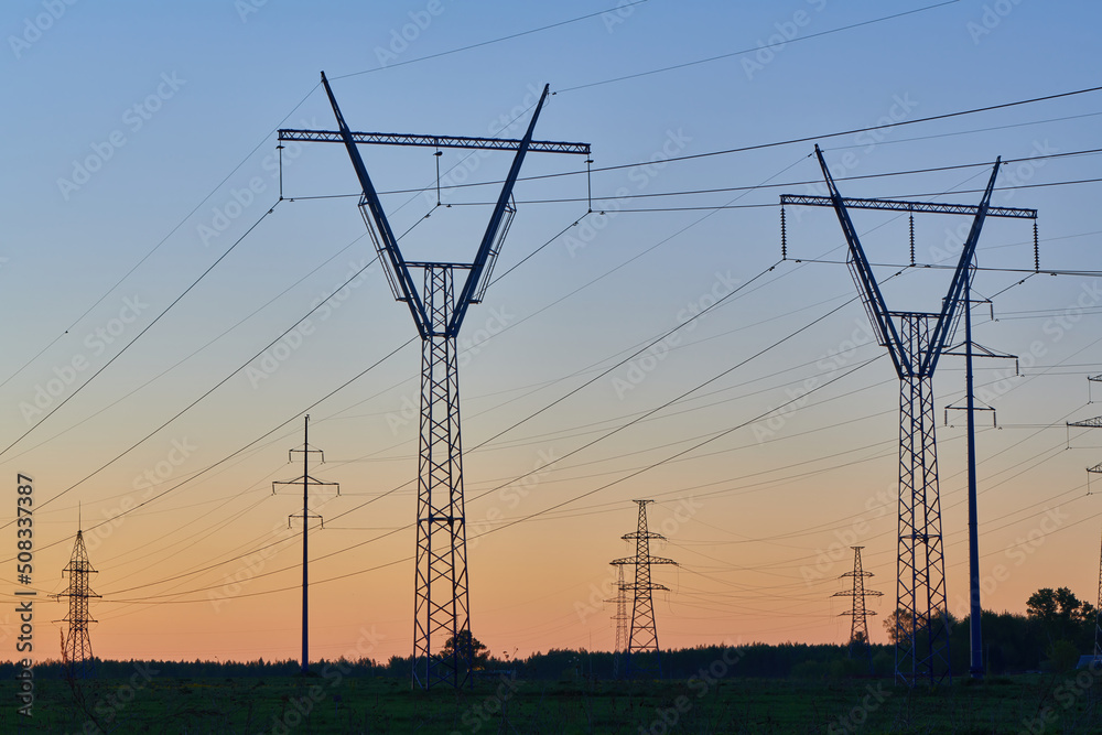 High voltage electricity towers in field at sunset and clear blue sky. Dark silhouettes of repeating power lines on orange sunrise. Electricity generation, transmission, and distribution network