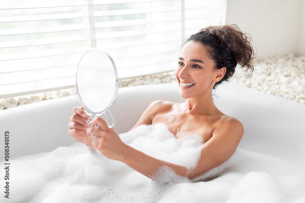 Sexy young woman looking at herself in mirror while taking hot bubble bath at home, copy space Sexy young woman looking at herself in mirror while taking hot bubble bath at home, copy space