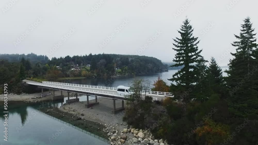Aerial view of the bridge and road crossing Moreno Lake in an early cloudy morning with fog. The forest and sky reflection in the water surface. The cars driving along the highway. Aerial view of the bridge and road crossing Moreno Lake in an early cloudy morning with fog. The forest and sky reflection in the water surface. The cars driving along the highway.