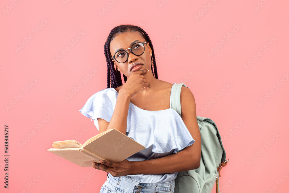 Pensive black female student with backpack holding open textbook, thinking over assignment, studying for exam Pensive black female student with backpack holding open textbook, thinking over assignment, studying for exam