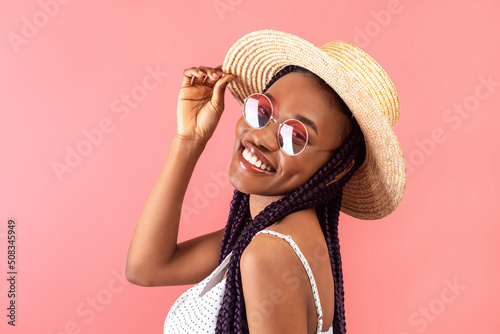 Cheerful black lady with braids wearing stylish dress and sunglasses, touching straw hat and smiling at camera