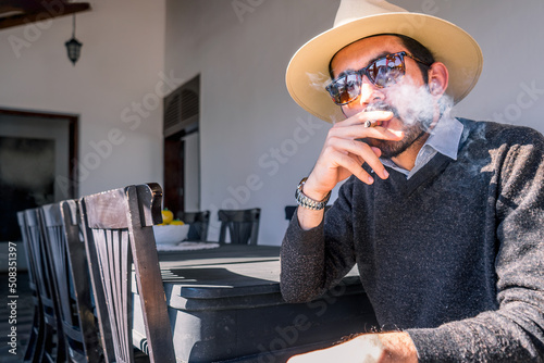 Young Nicaraguan man wearing a sweater hat and sunglasses smoking a cigar in the living room of a colonial-style mansion in Managua city