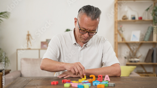 Korean male pensioner with Alzheimer’s disease trying to improve memory by doing rehab exercise for brain at home. He murmurs to himself while looking and counting the letter blocks
