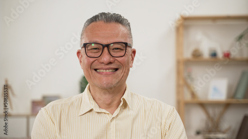 shoulder shot portrait of a cheerful Chinese elderly male in casual wear smiling at the camera in a modern bright home interior