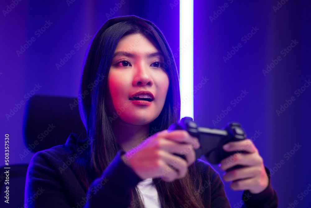 Playing video game. Young asian pretty woman sitting on chair holding joystick in living room. Happy female Professional Streamer chinese wearing hoodie playing game online in dark room neon light. Playing video game. Young asian pretty woman sitting on chair holding joystick in living room. Happy female Professional Streamer chinese wearing hoodie playing game online in dark room neon light.